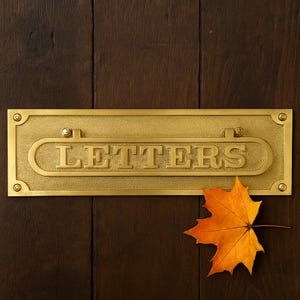 May include: A rectangular brass mailbox slot with the word "LETTERS" embossed on it. The mailbox slot is mounted on a dark brown wooden door. An orange and yellow maple leaf rests on the door.