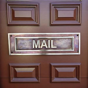 Pode incluir: Close-up de uma porta de madeira castanha com uma ranhura de correio de latão. A palavra "MAIL" está em letras maiúsculas. Acima e abaixo da ranhura estão dois painéis decorativos retangulares.