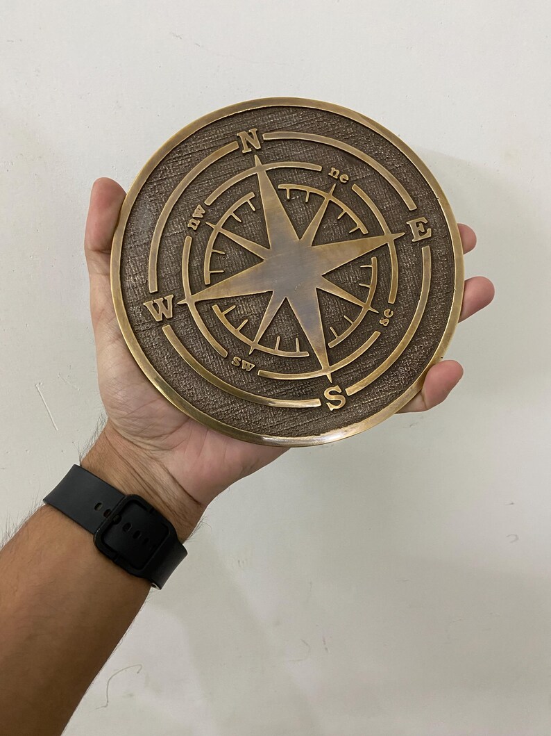 May include: A round, bronze-colored compass rose with directional markings (N, S, E, W, NE, NW, SE, SW) and a central star design. The compass is held in a hand, showcasing its size and detail.