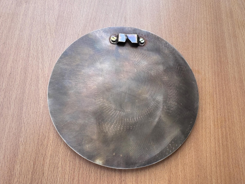 May include: A circular, metallic wall hanging with a brushed finish. The back features a metal bracket for hanging, secured with two screws. The piece has a vintage or industrial aesthetic.