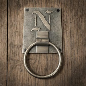 May include: A rectangular metal door knocker with a brushed silver finish featuring a large circular ring and a prominent letter N embossed on the top plate. The hardware is secured with four visible screws at the corners of the mounting plate.