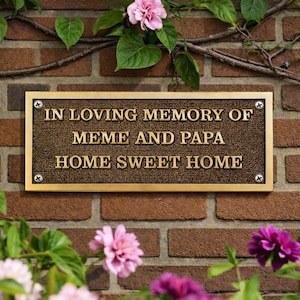 May include: A bronze plaque with the inscription "IN LOVING MEMORY OF MEME AND PAPA HOME SWEET HOME" is mounted on a brick wall. The plaque is surrounded by flowers and greenery, including pink and purple blooms.