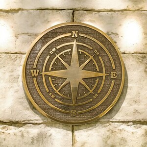 May include: A round, bronze-colored compass rose with directional markings (N, S, E, W, NE, NW, SE, SW) and a central eight-pointed star. The compass is mounted on a textured, circular background and set against a light-colored brick wall.