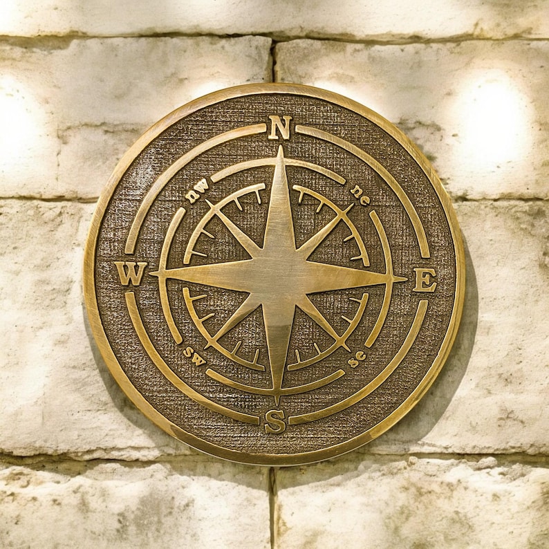 May include: A round, bronze-colored compass rose with directional markings (N, S, E, W, NE, NW, SE, SW) and a central eight-pointed star. The compass is mounted on a textured, circular background and set against a light-colored brick wall.
