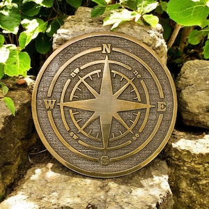 May include: A bronze-colored compass rose plaque with directional indicators (N, S, E, W) and a starburst design. The plaque is circular and has a textured background. The image is set against a backdrop of rocks and green foliage.