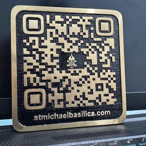 May include: A square, gold-colored QR code on a black background. The QR code is surrounded by a gold border and features the text "stmichaelbasilica.com". The image includes a small emblem in the center of the QR code.