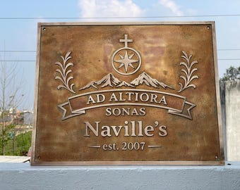 May include: A rectangular bronze-toned metal plaque featuring a raised design of a cross above a compass star, mountain peaks, and decorative sprigs. The text reads AD ALTIORA SONAS, Naville's, and est. 2007 on a banner and centered lines.