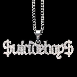 Puede incluir: Collar de cadena de plata con un colgante que dice "$WAVYBOYS$" en una fuente gótica, con cada letra cubierta de pequeños diamantes de imitación transparentes.