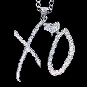 May include: A silver-toned pendant necklace featuring the letters XO in a stylized script font, fully encrusted with small clear stones. A small heart-shaped charm with matching stones is attached to the top of the letter O, suspended from a chain.