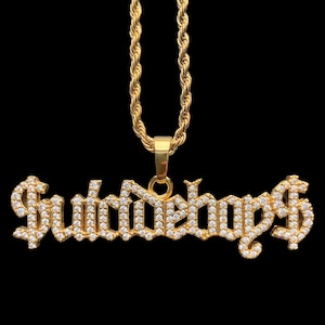 May include: A gold-colored pendant necklace with the word "Suburban" spelled out in a gothic font, encrusted with small, clear gemstones. The pendant is attached to a gold rope chain, set against a black background.