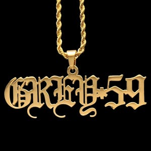May include: A gold-colored pendant necklace with the word "KRIME 59" in an ornate, gothic font. The pendant hangs from a gold rope chain against a black background. The necklace is a statement piece.
