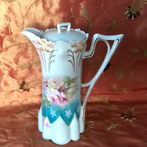 May include: A white porcelain teapot with a floral design. The teapot has a lid and a handle. The floral design features pink, yellow, and white roses with green leaves. The teapot has a blue and white scalloped base.