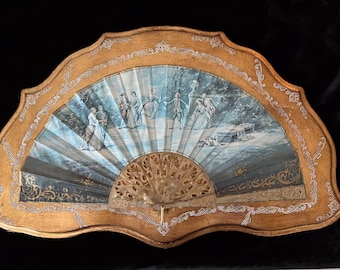 May include: A decorative fan with a gold frame and a painted scene of people in a garden. The fan is closed and the handle is visible.