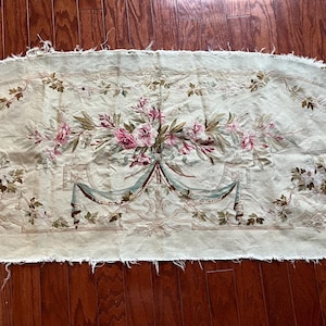 May include: A rectangular piece of fabric with a floral design. The fabric is a light green color with a distressed look. The floral design features pink flowers, green leaves, and blue ribbons. The edges of the fabric are unfinished.