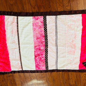 May include: A rectangular baby blanket with a brown satin border. The blanket features vertical stripes of bright pink, white, and pink patterned fabric. Decorative ribbons in brown and white with polka dots and stripes are sewn into the blanket.