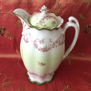 May include: A white porcelain creamer with a green and gold trim and a floral design. The creamer has a lid with a floral design and a scalloped edge.