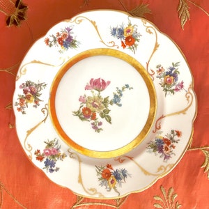 May include: A white porcelain plate with a gold rim and floral designs. The plate has a scalloped edge and features a variety of flowers in shades of pink, yellow, blue, and red.