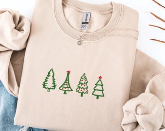 May include: A beige sweatshirt with embroidered green Christmas trees, each topped with a red star. A delicate silver necklace with a sun-shaped pendant rests on the sweatshirt. The sweatshirt is folded, with a portion of denim jeans visible.