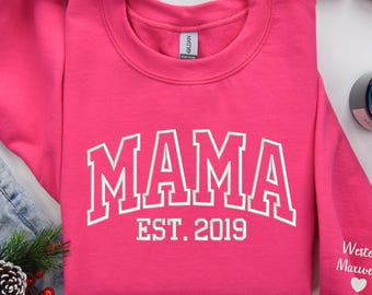 May include: A bright pink sweatshirt with the word "MAMA" in white, collegiate-style lettering. Below the word "MAMA" is the text "EST. 2019". The sleeve has the text "Wester Maxwell" and a heart symbol.