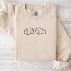 May include: A beige sweatshirt with a black embroidered design of five ghosts and bats. The ghosts have black eyes and mouths and are in different poses. The bats are orange and purple.