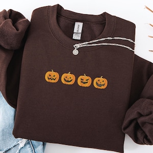 Op de afbeelding: Een donkerbruine sweatshirt met vier geborduurde oranje pompoenen over de borst. Een zilveren ketting met een kleine hanger is over de sweatshirt gedrapeerd. De sweatshirt heeft een ronde hals en lange mouwen.