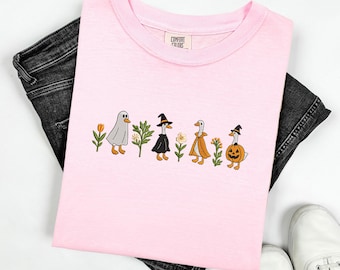 May include: A light pink t-shirt with an embroidered Halloween design featuring geese dressed as a ghost, witch, and pumpkin. The shirt is folded over black denim jeans and white sneakers.