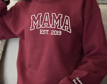 May include: A maroon sweatshirt with the word "MAMA" in white, outlined block letters. Below the word "MAMA" is the text "EST. 2019". The sleeve has the name "Weston Maxwell" and a heart symbol.