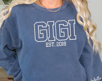 May include: A blue sweatshirt with the word "GIGI" in white block letters, with "EST. 2018" below. The sleeve has the name "Isabella Noah" and a heart. The sweatshirt is a solid blue color.