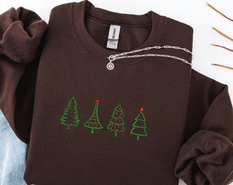 May include: A dark brown sweatshirt with four embroidered green Christmas trees, each with a unique design. The trees have red star accents. A silver necklace with a sunburst pendant rests on the sweatshirt.