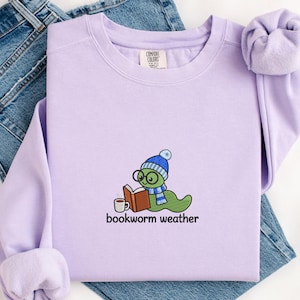 Può includere: Una felpa girocollo color viola chiaro con un ricamo raffigurante un verme verde che indossa un berretto e una sciarpa a righe blu e bianche, intento a leggere un libro aperto con una tazza di caffè accanto e la scritta bookworm weather.