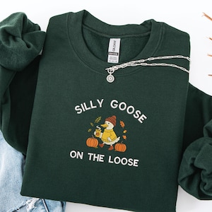 Peut inclure: Un sweat-shirt vert foncé avec l'inscription blanche "SILLY GOOSE ON THE LOOSE". Le motif représente une oie de dessin animé avec une tasse de café, des citrouilles et des feuilles d'automne. Un collier argenté avec un pendentif fleur est posé sur le sweat-shirt.