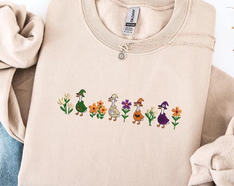 May include: Beige sweatshirt with embroidered geese wearing witch hats and walking among flowers. The design includes green, orange, purple, and white floral accents. A delicate silver necklace with a sun pendant is draped on the sweatshirt.