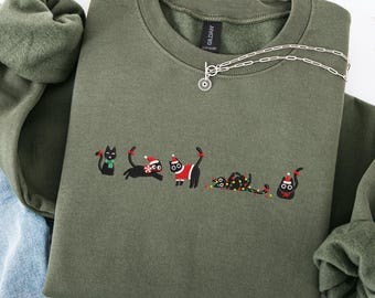 May include: An olive green sweatshirt featuring an embroidered design of black cats in festive holiday attire. A silver necklace with a circular pendant is draped on the sweatshirt. The sweatshirt is folded with a green fleece scarf and blue jeans.