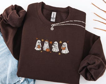 May include: A dark brown sweatshirt featuring an embroidered design of four cartoon ghosts, each with a pumpkin, pumpkin hat, coffee cup, and book. A silver necklace with a sun pendant is on the sweatshirt.