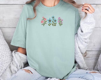 May include: A light blue t-shirt with a row of embroidered flowers in pink, blue, and green.