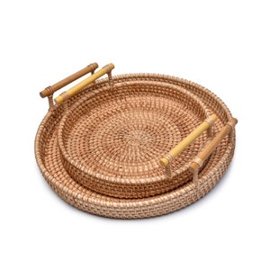May include: Two round woven rattan trays, one nested inside the other. Each tray has a light brown woven pattern and wooden handles. The handles are a light brown colour with a yellow accent.