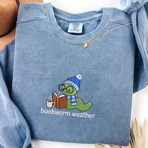 Op de afbeelding: Een blauwe sweater met ronde hals en een geborduurde afbeelding van een groene worm met een blauw-wit gestreepte muts, sjaal en een ronde bril. De worm leest een open boek naast een dampende mok. De tekst op de sweater luidt bookworm weather.