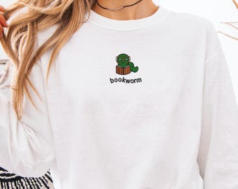 May include: White sweatshirt with a small embroidered design of a green worm reading a book. The word "bookworm" is written in black below the design. The sweatshirt is made of a soft material.