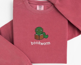 May include: A maroon Comfort Colors sweatshirt featuring a cartoon bookworm wearing glasses and reading a book. The word "bookworm" is embroidered below the graphic. The sweatshirt has long sleeves and a crew neck.