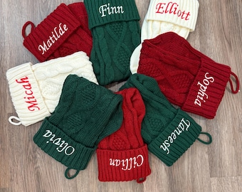 Op de afbeelding: Een verzameling gepersonaliseerde kerstsokken in rood, groen en crème. Elke sok heeft een kabelgebreid patroon en een manchet met een naam geborduurd in een contrasterende kleur. De namen zijn Matilde, Finn, Elliott, Micah, Olivia, Cillian, Sophia en Taneesh.