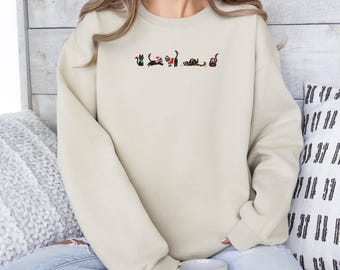 May include: A cream-coloured sweatshirt with an embroidered design of cats in different poses. The cats are black with red and green details, creating a festive look. The sweatshirt is made of a soft material.