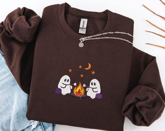 May include: A dark brown sweatshirt with an embroidered design of two white ghosts roasting marshmallows over a campfire. The design includes a crescent moon and stars. A silver necklace is draped across the sweatshirt.