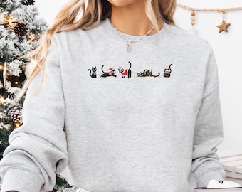 May include: A pale grey sweatshirt with a series of black cat illustrations, each decorated with Christmas-themed elements. The cats are shown in different poses, some wearing Santa hats and others with festive decorations. The design is simple and whimsical.