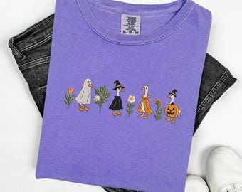 May include: A purple Comfort Colors t-shirt featuring an embroidered design of geese dressed in Halloween costumes. The geese are depicted as a ghost, witch, and pumpkin, with flowers and plants. The shirt is folded on top of black jeans and white sneakers.