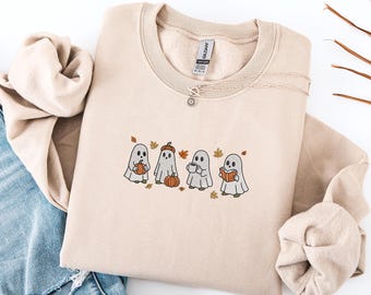 May include: Beige crew neck sweatshirt featuring an embroidered design of four cartoon ghosts, each with a different autumn-themed item: a pumpkin, a pumpkin hat, a coffee cup, and a book. The sweatshirt is paired with a silver necklace and blue jeans.
