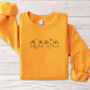 May include: A bright orange sweatshirt with a cute design of four ghosts and bats. The ghosts are white with black eyes and mouths. The bats are black with purple wings.