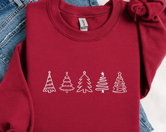 May include: A burgundy sweatshirt featuring a white line drawing of five different Christmas trees. The sweatshirt is made of a soft material and is folded on top of a pair of blue jeans.