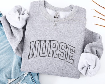 Puede incluir: Una sudadera gris con la palabra "NURSE" impresa en negro en la parte delantera. La sudadera está doblada y colocada sobre una superficie blanca.