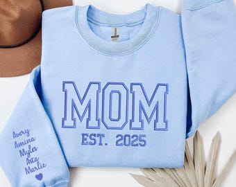 May include: Light blue sweatshirt with the word "MOM" in blue outlined letters and the text "EST. 2025" below. The left sleeve has the names Avery, Amina, Myla, Ava, and Marlie written in white.