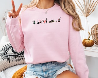 May include: A pale pink sweatshirt with a series of black cat illustrations. Some cats are depicted with festive Christmas decorations. The sweatshirt has a crew neck and long sleeves.